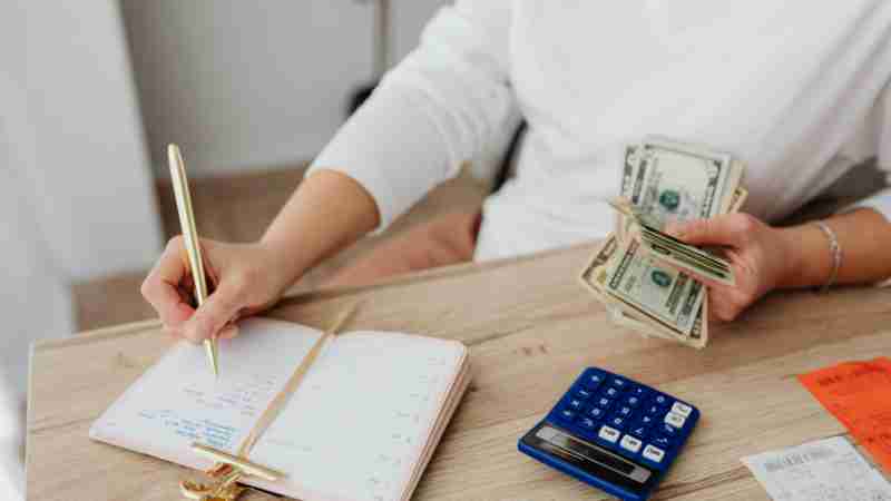 Adult holding cash and writing in planner while using a calculator at home., tags: ihr einen finanzplan - pexel